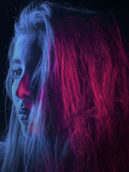 A woman's profile is captured with vibrant lighting. Her face is partially illuminated with blue and pink light, creating a striking contrast against the dark background. The colors blend into her long hair, giving a dramatic effect.