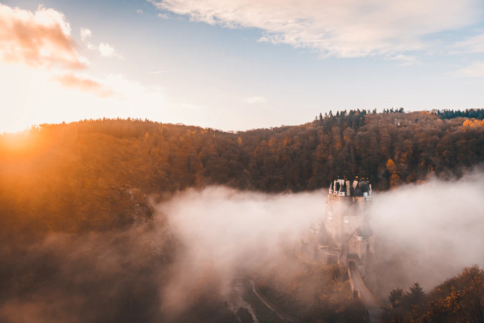 The image depicts a serene landscape with a castle-like structure nestled amidst a foggy hillside, surrounded by trees and hills. The sky above is painted in shades of orange and yellow, suggesting either sunrise or sunset. The fog obscures the view slightly but does not diminish the overall beauty of the scene.