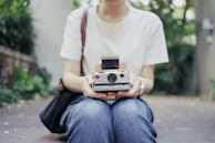 A person sitting cross-legged outdoors while holding a vintage instant camera. The individual is dressed in casual jeans and a white t-shirt. The background features greenery and a blurred outdoor setting, suggesting a relaxed and leisurely environment.