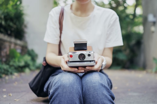 A person sitting cross-legged outdoors while holding a vintage instant camera. The individual is dressed in casual jeans and a white t-shirt. The background features greenery and a blurred outdoor setting, suggesting a relaxed and leisurely environment.