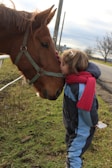 A child dressed in winter clothing and a red scarf is gently kissing the nose of a brown horse with a harness. They are standing on a grassy area beside a rural road, with leafless trees and a cloudy sky in the background, creating a serene countryside setting.