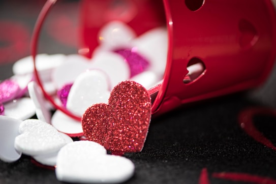 A collection of glittery heart-shaped decorations is scattered around and spilling out of a small red bucket. The hearts are predominantly red, white, and pink, with a glossy, sparkly texture. The red bucket has heart-shaped cutouts and is lying on its side.