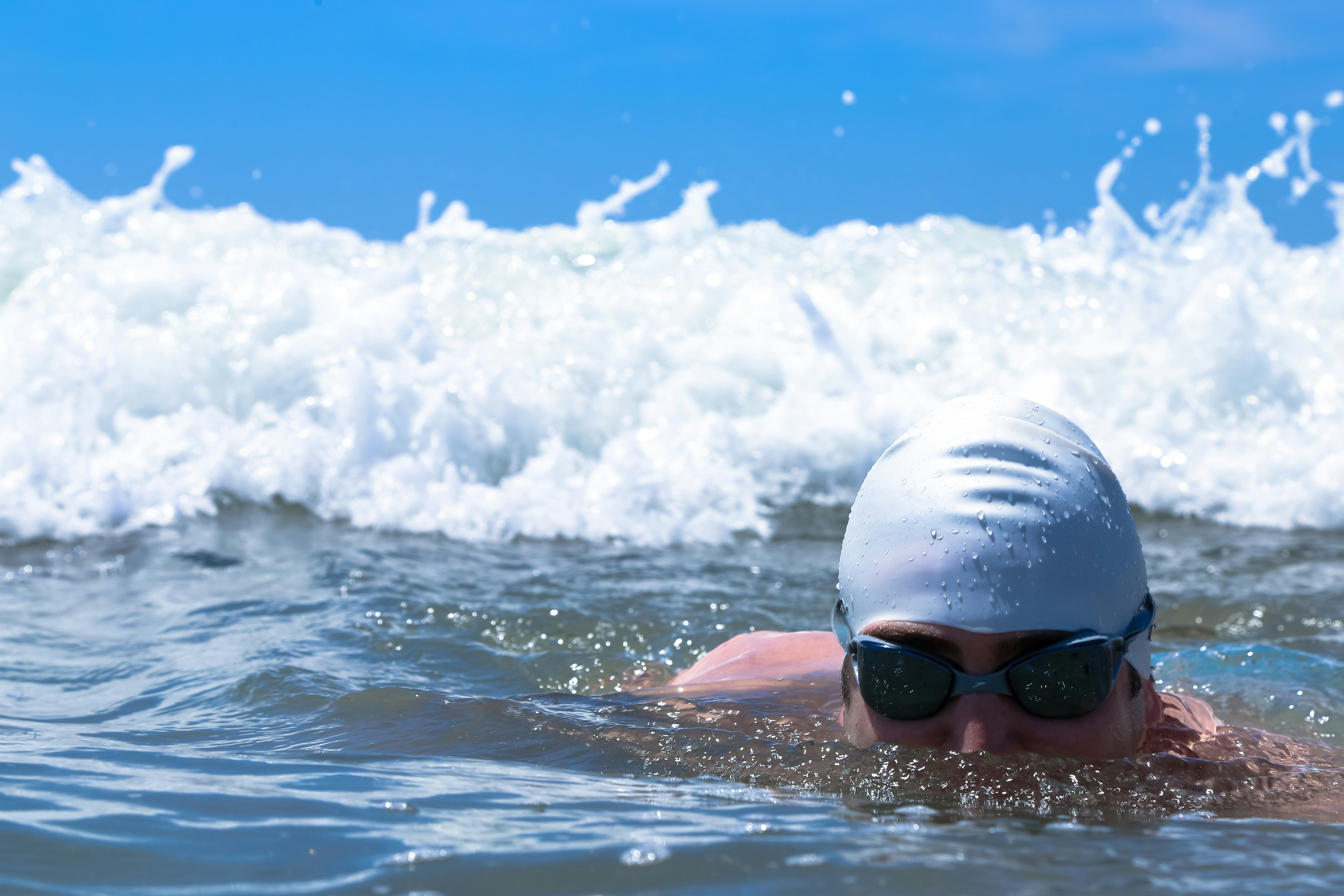 A swimmer wearing a white swim cap and goggles emerges from the water, surrounded by ocean waves against a bright blue sky.