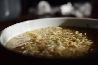 A close-up view of a bowl filled with instant noodles in broth, with soft lighting casting shadows and highlighting the texture of the noodles.
