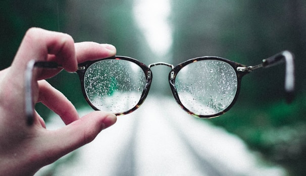 A hand holds a pair of eyeglasses with raindrops on the lenses, creating a clear view through the glasses against an out-of-focus background of a path in a forested area.