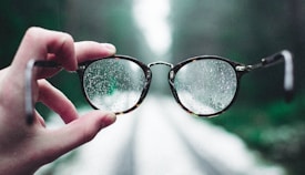 A hand holds a pair of eyeglasses with raindrops on the lenses, creating a clear view through the glasses against an out-of-focus background of a path in a forested area.