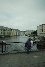 A picturesque waterfront scene with a canal lined by colorful buildings with steep, slate roofs. A bridge in the foreground has ornate, wrought-iron railings where a person stands looking at a map. Several boats are moored along the water, and the sky is overcast.