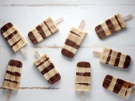 Eight chocolate and vanilla striped popsicles are scattered on a light wooden surface. The popsicles are arranged in various angles, displaying their distinct striped patterns. The wooden sticks and creamy textures contrast with the background.