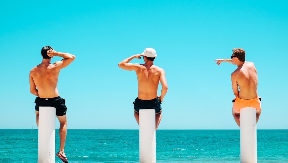 three men's sitting on white concrete bollard near body of water during daytime