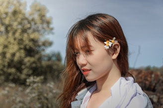 A young woman with long hair is outdoors, with daisies tucked behind her ear. She is looking off to the side, and the background features blurred greenery and a soft blue sky.