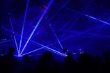 A dimly lit environment with numerous blue laser beams crisscrossing in various directions. Silhouettes of people are visible, suggesting a lively atmosphere, likely a party or concert.