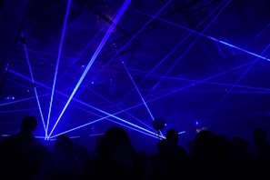 A dimly lit environment with numerous blue laser beams crisscrossing in various directions. Silhouettes of people are visible, suggesting a lively atmosphere, likely a party or concert.