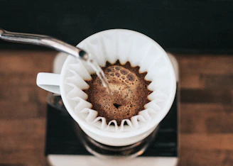Close-up image of a kettle pouring hot water over coffee grounds in a V60 dripper, with coffee beans and filters nearby.