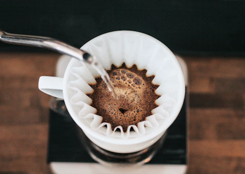 Close-up image of a kettle pouring hot water over coffee grounds in a V60 dripper, with coffee beans and filters nearby.
