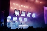 A vibrant concert scene with large LED panels displaying bright, vertical white light patterns hanging from above. The stage is filled with smoke, creating a dramatic effect illuminated by the purple and blue lighting. The audience is visible in silhouette with raised hands, and there are performers on a lit platform surrounded by equipment.