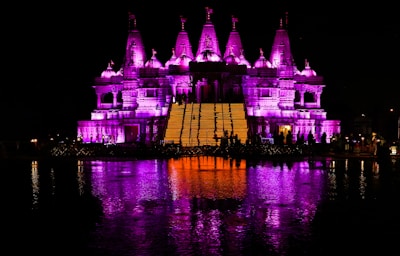 A beautifully illuminated temple with intricate architecture is lit with vibrant purple lighting. The temple reflects in a body of water in the foreground, adding to the symmetrical aesthetic. Silhouettes of people are visible near the temple, enhancing the sense of scale and activity.
