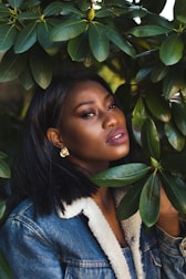 A woman with short black hair wearing a denim jacket with a shearling collar and gold earrings is partially obscured by green leaves. She gazes softly at the camera, with a background filled with foliage.
