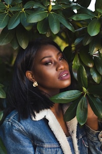 A woman with short black hair wearing a denim jacket with a shearling collar and gold earrings is partially obscured by green leaves. She gazes softly at the camera, with a background filled with foliage.