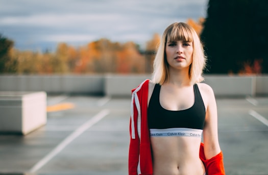 A young woman with blonde hair is standing outdoors. She is wearing a black sports bra with a visible Calvin Klein logo band and a red jacket draped over her shoulders. The background is an unfocused urban setting with autumn-colored trees.