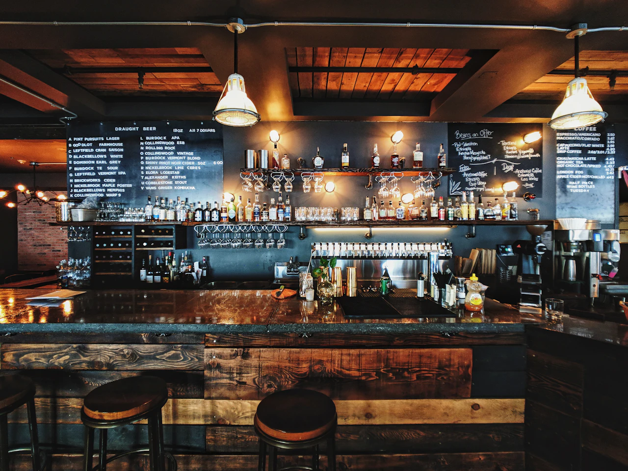 The wooden bar counter at night, bottles lined up under chalkboard menus