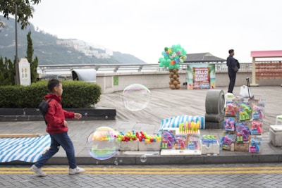 A young boy wearing a red jacket is playfully interacting with large, colorful bubbles. He appears on a street with a vendor's stall displaying an array of colorful toys and bubble wands. In the background, there is a sign for Stanley Market and decorations made from balloons. The setting is outdoors, and further back, a man stands near some railings and a scenic view of hills and distant buildings.