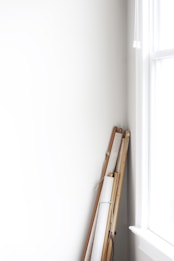 Several rolled canvases or scrolls with wooden poles are leaning against a white wall near a bright window. The scene is minimalistic with soft natural lighting.