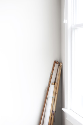Several rolled canvases or scrolls with wooden poles are leaning against a white wall near a bright window. The scene is minimalistic with soft natural lighting.