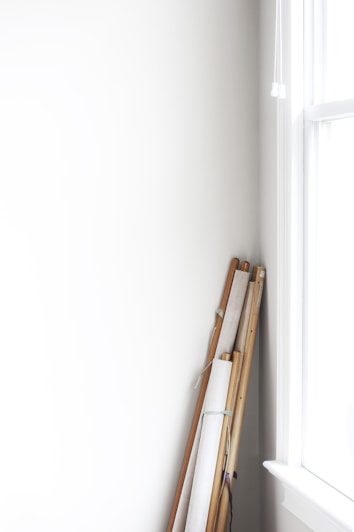 Several rolled canvases or scrolls with wooden poles are leaning against a white wall near a bright window. The scene is minimalistic with soft natural lighting.