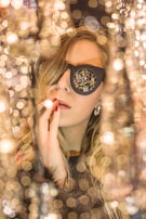 A person wearing black sunglasses surrounded by a dazzling display of lights, creating a bokeh effect. The scene is glamorous and filled with reflections, highlighting the person's features and creating an artistic atmosphere.