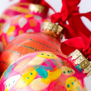 Three colorful ornaments adorned with abstract patterns and vibrant colors like pink, orange, and yellow, each topped with a metallic gold cap and tied with a bright red ribbon.