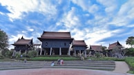 A traditional Thai architectural complex with multiple gabled roofs, elevated platforms, and dark wooden structures set against a vibrant blue sky. The area is surrounded by greenery, including shrubs and trees, with people sitting and walking on the steps and pathways leading to the buildings.