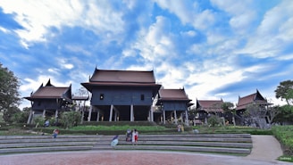 A traditional Thai architectural complex with multiple gabled roofs, elevated platforms, and dark wooden structures set against a vibrant blue sky. The area is surrounded by greenery, including shrubs and trees, with people sitting and walking on the steps and pathways leading to the buildings.