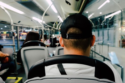 A view from the interior of a bus, focusing on the back of a passenger wearing a black cap. The bus is modern, with sleek seating and overhead lighting. Other passengers are seated throughout the bus, and there is a large glass window on the right side, hinting at an urban setting outside.