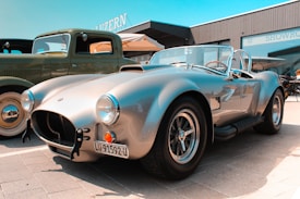 A silver vintage sports car with a sleek design is parked beside an older green vehicle, possibly a classic truck. The sports car has a prominent front grille and round headlights. Its shiny exterior reflects the sunlight, highlighting its curves. The background includes a modern gray building with a sign mentioning 'SHOWROOM'.