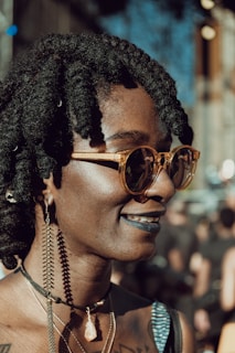 A person with braided hair wears stylish round sunglasses, large hoop earrings, and layered necklaces with a distinctive stone pendant. The smile conveys a confident and relaxed demeanor, enhanced by dark lipstick. The background suggests an outdoor setting with blurred figures and structures.