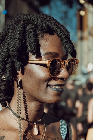 A person with braided hair wears stylish round sunglasses, large hoop earrings, and layered necklaces with a distinctive stone pendant. The smile conveys a confident and relaxed demeanor, enhanced by dark lipstick. The background suggests an outdoor setting with blurred figures and structures.