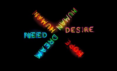 Neon words in various colors arranged in a cross pattern against a black background. The words 'HUMAN', 'DESIRE', 'NEED', 'DREAM', and 'HOPE' are displayed, each in its own vibrant hue.
