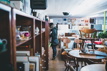 A person is standing in an indoor space filled with various second-hand furniture and household items. Shelves are lined with miscellaneous objects such as teapots and picture frames. Wooden tables, chairs, and other furniture pieces are spread throughout the area in a somewhat cluttered arrangement. The space has a cozy, somewhat nostalgic atmosphere typical of a thrift store or antique shop.
