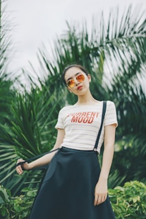 A person stands confidently in front of lush green foliage, wearing mirrored sunglasses and a striped shirt with the text 'CURRENT MOOD'. The attire includes a dark skirt held by suspenders. The background consists of vibrant palm leaves, adding a tropical vibe.