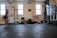 A person is performing a fitness exercise using battle ropes in a gym. The gym environment includes various types of equipment such as weights, resistance bands, and a squat rack. The floor is covered with black mats, and there are large windows allowing natural light. The walls have motivational text and there is a framed picture.