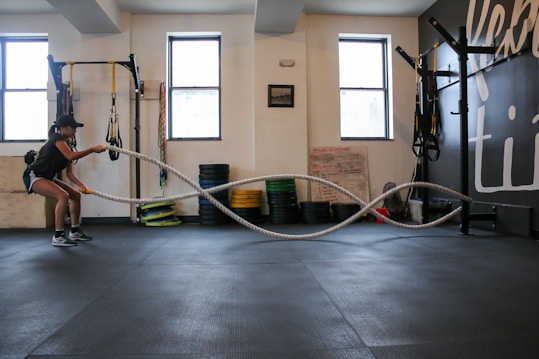 A person is performing a fitness exercise using battle ropes in a gym. The gym environment includes various types of equipment such as weights, resistance bands, and a squat rack. The floor is covered with black mats, and there are large windows allowing natural light. The walls have motivational text and there is a framed picture.