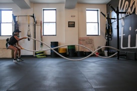 A person is performing a fitness exercise using battle ropes in a gym. The gym environment includes various types of equipment such as weights, resistance bands, and a squat rack. The floor is covered with black mats, and there are large windows allowing natural light. The walls have motivational text and there is a framed picture.