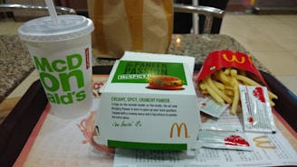 A McDonald's meal consisting of a large drink cup, a box containing a McSpicy Paneer burger, and a packet of fries partially opened. The items are placed on a tray with table condiments like ketchup packets. The setting is a fast-food restaurant with a modern interior design.