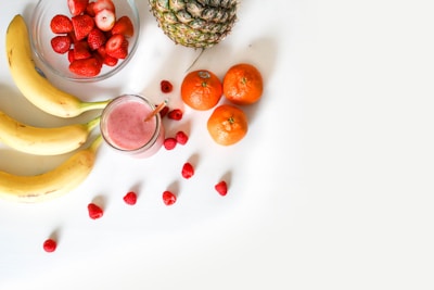 A mix of fresh fruits elegantly arranged on a white background. Three bananas, a glass of pink smoothie, several strawberries in a glass bowl, a whole pineapple, three oranges, and scattered raspberries are present. The smoothie is positioned centrally accompanied by a straw.