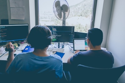 Two individuals are seated at a workstation with multiple monitors displaying lines of code. Both appear to be focused on their tasks, with one person using a laptop and the other with a desktop setup. A large window in the background reveals a scenic outdoor view. The desk is cluttered with various items including a fan, a headset, and some papers.