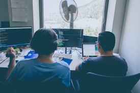 Two individuals are seated at a workstation with multiple monitors displaying lines of code. Both appear to be focused on their tasks, with one person using a laptop and the other with a desktop setup. A large window in the background reveals a scenic outdoor view. The desk is cluttered with various items including a fan, a headset, and some papers.