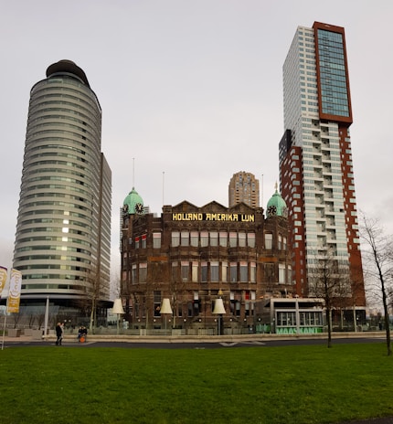 A historic building with the sign 'Holland Amerika Lijn' is flanked by two modern skyscrapers. The architecture contrasts old and new styles, with the older building featuring ornate details and the skyscrapers showing sleek, contemporary designs. In the foreground, there is a green lawn, and a few people are walking nearby.