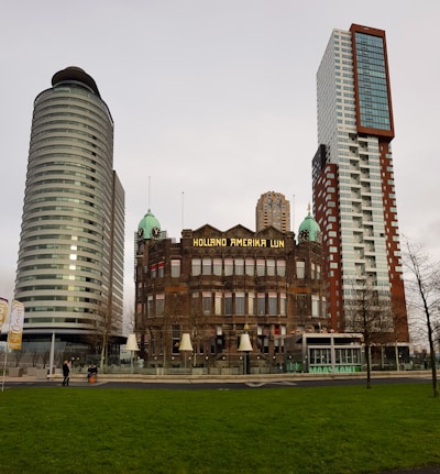 A historic building with the sign 'Holland Amerika Lijn' is flanked by two modern skyscrapers. The architecture contrasts old and new styles, with the older building featuring ornate details and the skyscrapers showing sleek, contemporary designs. In the foreground, there is a green lawn, and a few people are walking nearby.