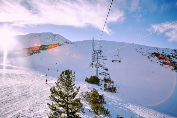 A chairlift climbing a wide groomed piste with skiers and scattered pines under low winter sun