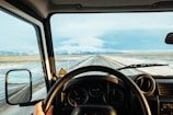 A driver’s perspective from inside a moving vehicle, showing a vast road stretching into the distance bordered by snow-covered plains and a mountain range under a partly cloudy sky. The dashboard and steering wheel are visible, indicating a focus on travel or journey.